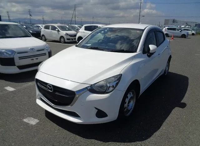 A White Mazda Compact Car Is Parked In An Outdoor Lot Surrounded By Other Vehicles Under A Partly Cloudy Sky. The Car, Possibly On Offer By Mundiya, Faces Slightly Towards The Camera From The Front-Right Side, Suggesting It'S Part Of A Lively Car Sale Event.