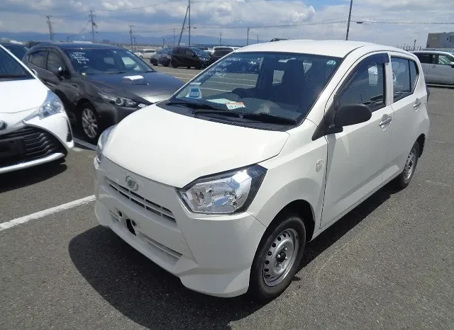 A White Compact Car, Possibly A Recent Import For Sale, Is Parked In A Bustling Lot On A Clear Day. Surrounded By Other Vehicles Under The Kenyan Sky, Power Lines Stretch Across The Cloudy Background.