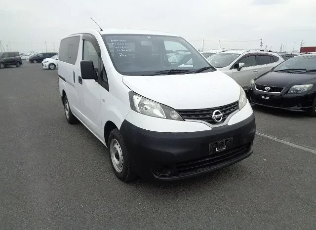 A White Nissan Van, Possibly Part Of A Car Sale In Kenya, Is Parked Among Other Vehicles. The Van Faces Slightly Left, Captured From A Front Side Angle Under An Overcast Sky On Paved Ground.